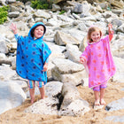 Two children on a beach in hooded Beach Towel Ponchos with star prints, waving to the camera.